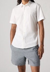 Weißes kurzärmliges Hemd mit Knopfkragen, kombiniert mit blau-weiß gestreiften Shorts, die eine lockere Passform und glatte Stofftextur aufweisen.