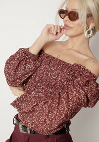 Bordowy kwiatowy top bez ramiączek z bufiastymi rękawami i elastycznym dołem; w zestawie z spodniami z wysokim stanem i oversize'owymi okularami przeciwsłonecznymi.