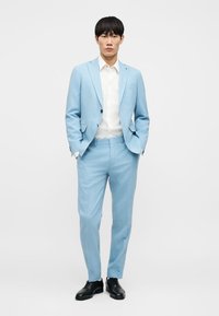 Costume bleu clair en tissu texturé à coupe ajustée, accompagné d'une chemise blanche et de chaussures noires, présentant des lignes épurées et un design minimaliste.