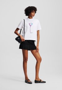Witte katoenen t-shirt met een kleurrijke bloemenafbeelding, gecombineerd met een zwarte geplooide rok en platte schoenen met witte stippen, en een zwarte schoudertas.