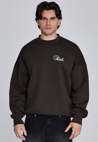 Brązowy, oversize'owy sweatshirt z okrągłym dekoltem, opadającymi ramionami i ściągaczami. Posiada biały napis z logo na lewej części klatki piersiowej.