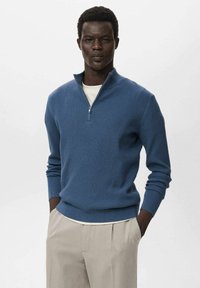Modra rebrasta pulover z ovratnikom na zadrgo, dolgimi rokavi in prilegajočim se dizajnom. V kombinaciji s bež hlačami, prikazan na enostavni podlagi.