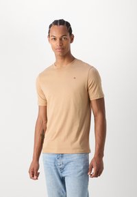 Camiseta de manga corta beige hecha de una tela suave, con cuello redondo y un pequeño logotipo en el pecho, combinada con jeans de color azul claro.