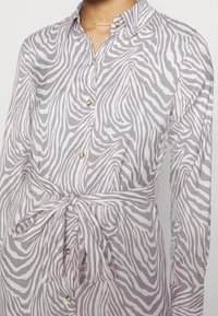 Lehká průsvitná košile s fialovo-bílým zebrovým vzorem, s knoflíkovou légou a detailem v podobě zavazování v pase. Dlouh é rukávy s knoflíkovými akcenty.