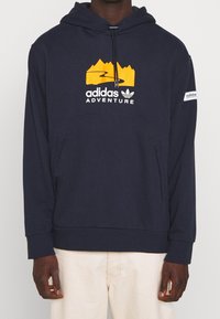 Tmavě modrá mikina s kapucí z měkké látky, která má horskou grafiku a text "adidas ADVENTURE". Obsahuje přední kapsu a ribované manžety.