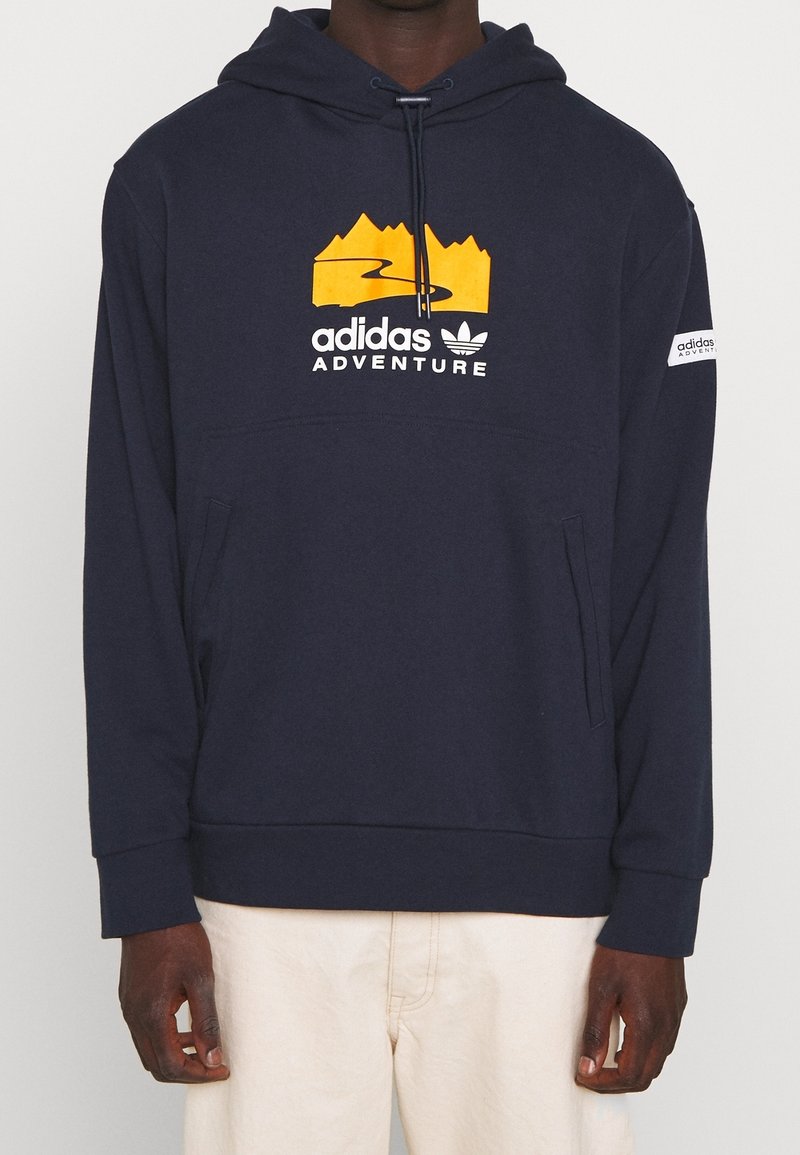 Tmavě modrá mikina s kapucí z měkké látky, která má horskou grafiku a text "adidas ADVENTURE". Obsahuje přední kapsu a ribované manžety.