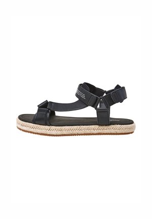 Musta nahkasaapas, jossa on espadrillo-tyylinen kudottu ina, säädettävät hihnat ja teksturoitu pohja mukavuutta varten.