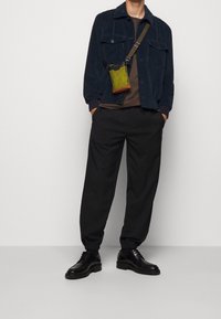 Mężczyzna w granatowej kurtce z kordurowej tkaniny, brązowej koszuli, czarnych zwężających się spodniach, czarnych skórzanych butach i zielonej torbie na ramię, z rękami w kieszeniach.