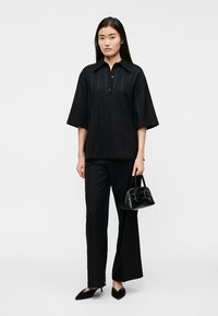 Donna che indossa pantaloni neri a gamba larga, camicia nera a maniche corte con colletto, tacchi neri appuntiti e tiene una piccola borsa nera.