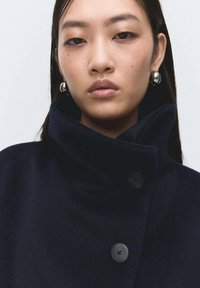 Abrigo de lana azul marino con un cuello alto, que presenta grandes botones negros y una textura suave. La modelo lleva pendientes de aro plateados.