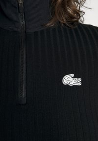 Gros plan d'un haut noir à côtes avec fermeture éclair et un logo crocodile blanc sur la poitrine, avec des cheveux bruns bouclés partiellement visibles.