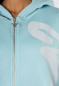 Felpa con zip azzurro chiaro realizzata in tessuto morbido, con una grande grafica bianca. Accessori in argento sulla zip, con una linguetta a forma di farfalla.
