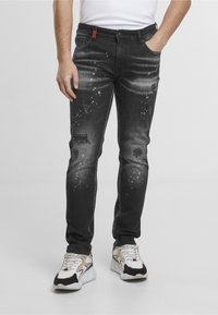 Homme portant un jean noir usé avec des éclaboussures de peinture blanche et des baskets blanches, une main dans la poche, sur un fond uni.