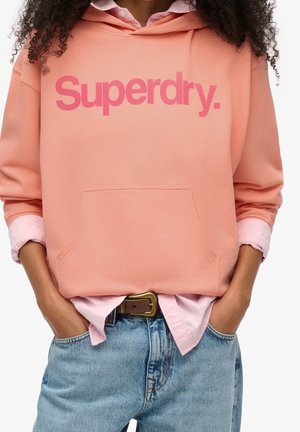 Persona vistiendo una sudadera con capucha Superdry color durazno sobre una camiseta rosa metida en unos vaqueros azules con cinturón marrón, con las manos en los bolsillos.