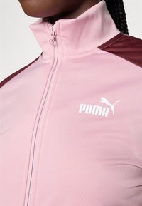 PUMA sporta jaka gaiši rozā krāsā ar augstu apkakli, ar bordo akcentiem un baltu logo priekšā, izgatavota no gludas, elastīgas auduma.