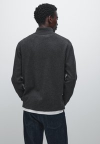 Dark gray wool blend pullover with a high collar and long sleeves, styled with a white layered shirt underneath and blue denim jeans.