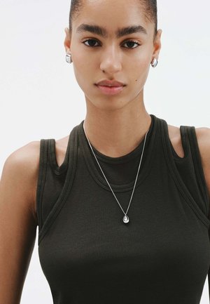 Joven mujer con una blusa sin mangas de color oscuro con detalles recortados, collar de medallón plateado en forma de lágrima y pendientes de aro plateados contra un fondo liso.