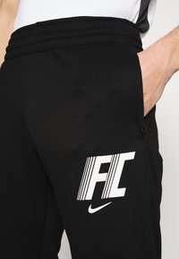 Zwarte sportbroek met een geribbelde tailleband, zijzak en een witte bedrukking met "FC" en het Nike-logo op het onderbeen.