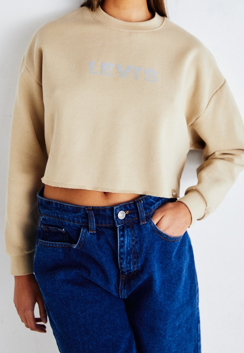 Personne portant un sweat-shirt beige coupé Levi's et un jean bleu taille haute avec une main dans la poche, sur fond blanc.