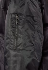 Černá látka bundy s hladkým povrchem, vybavená kapsou na boku se zipem a viditelnými zuby zipu. Jednoduchý, utilitární design.