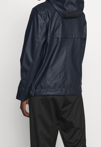 Marinblå vattentät jacka med huva, slät textur och avslappnad passform. Har ett bakstycke och långa ärmar med manschettdetaljer.