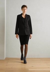 Blazer noir ajusté avec deux poches avant, associé à une jupe noire au genou et des collants noirs transparents, porté avec des bottines.