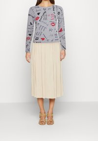 Haut gris à manches longues avec motifs graphiques rouges et noirs, associé à une jupe midi beige plissée, et des sandales à brides de couleur claire.