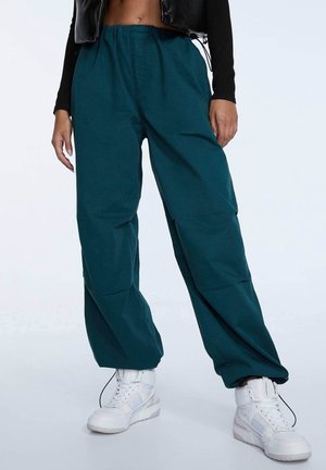 Persoon met turquoise wijde broek, witte hoge sneakers en een zwart cropped top met lange mouwen en een zwart vest.