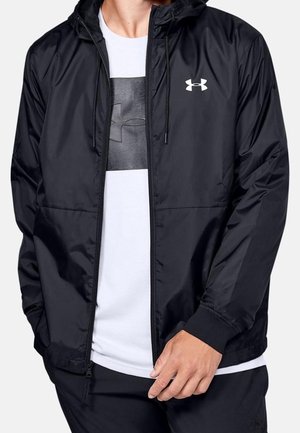 Mann trägt eine schwarze Under Armour Zip-up-Jacke über einem weißen T-Shirt mit Under Armour-Logo, Hände in den Taschen.