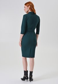 Figuurvormende teal jurk met driekwart mouwen, tailleband, rugopening en hoge kraag. Gecombineerd met zwarte enkellaarsjes met hardware-accenten.
