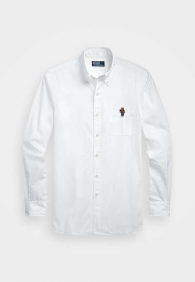 Balta garām piedurknēm poga-up krekls ar pogām uz leju, kabata uz krūtīm ar mazu izšūtu lāci, kas valkā svītrainu apģērbu.