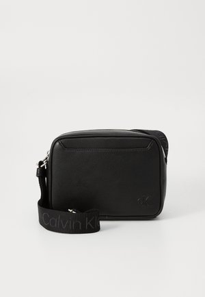 Sac bandoulière Calvin Klein noir en cuir texturé, forme rectangulaire, fermeture éclair et bandoulière ajustable en tissu brandé.