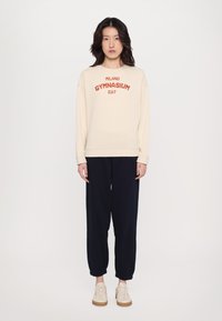 Beige spalvos crewneck džemperis su raudonu užrašu "MILANO GYMNASIUM", derinamas su tamsiai mėlynomis jogger kelnėmis ir šviesiais sportiniais bateliais. Ypatingas laisvas siluetas.