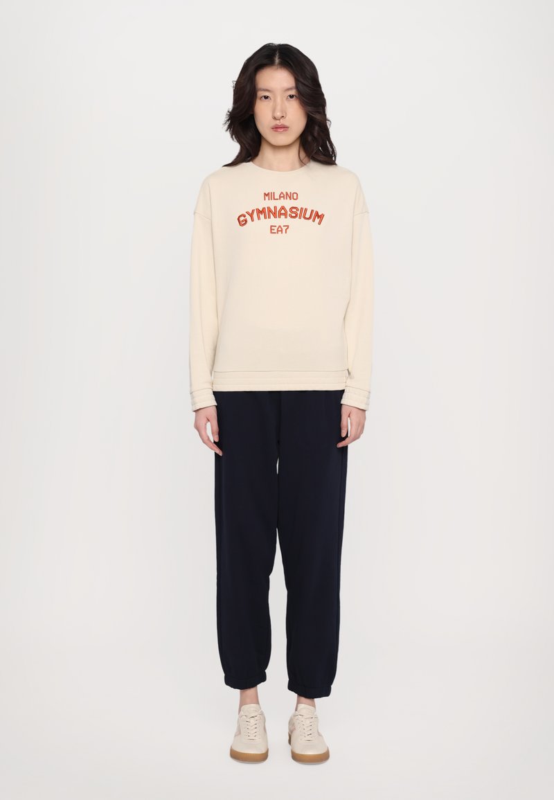 Beige spalvos crewneck džemperis su raudonu užrašu "MILANO GYMNASIUM", derinamas su tamsiai mėlynomis jogger kelnėmis ir šviesiais sportiniais bateliais. Ypatingas laisvas siluetas.