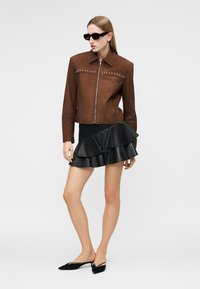 Γυναίκα που φοράει καφέ μπουφάν με φερμουάρ και μεταλλικά τρουκς, μαύρη πολυεπίπεδη φούστα με βολάν, μαύρα γυαλιά ηλίου και μαύρα mule με μυτερή μύτη, στέκεται μπροστά σε λευκό φόντο.