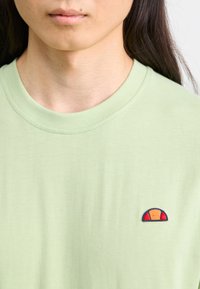 Light green cotton t-shirt featuring a small, embroidered patch with a red, orange, and navy design on the left chest area.