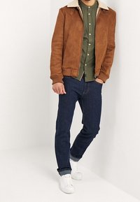 Mand iført en brun shearling-jakke, en grøn button-up skjorte, mørkeblå jeans og hvide sneakers, står op ad en hvid væg.