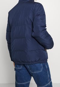 Marinblå puffjacka med hög krage, med kviltade paneler, elastisk nederkant och ärmslut, samt sidofickor med dragkedja. Slät, vattenavvisande material.