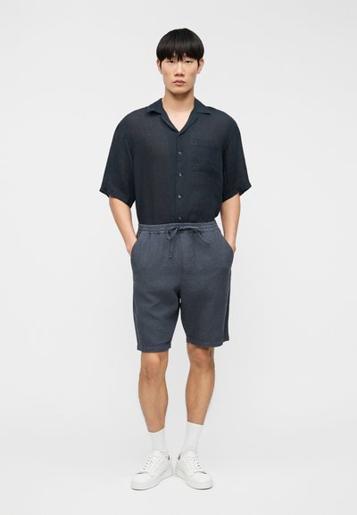 Homme debout les mains dans les poches, portant une chemise noire à manches courtes avec boutons, un short gris avec cordon de serrage, des chaussettes blanches et des baskets blanches.