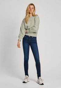 Sudadera verde claro con hombros caídos, combinada con jeans ajustados azul oscuro y zapatillas grises, con un diseño sencillo sin patrones.