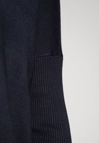 Marinblå tröja med ribbad struktur vid ärmsluten och sidorna, med ett mjukt, fint stickat tyg. Nätt närbild av ärmdetaljer.