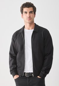 Vyras su tamsiais garbanotais plaukais, dėvintis juodą zip-up striukę, baltą marškinėlį ir juodas džinsus, su rankomis kišenėse, atsisukęs į priekį prieš paprastą foną.