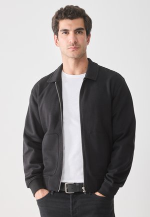 Man with dark curly hair wearing black zip-up jacket, white t-shirt, and black jeans with hands in pockets, facing forward against plain background.