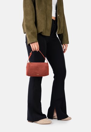 Person wearing olive green jacket, black flared pants, beige shoes, holding small rust-colored handbag with short strap.