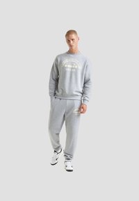 Felpa grigia con polsini a coste e scollatura arrotondata, caratterizzata da testo grafico bianco. Abbinata a pantaloni della tuta grigi coordinati e sneakers bianche.