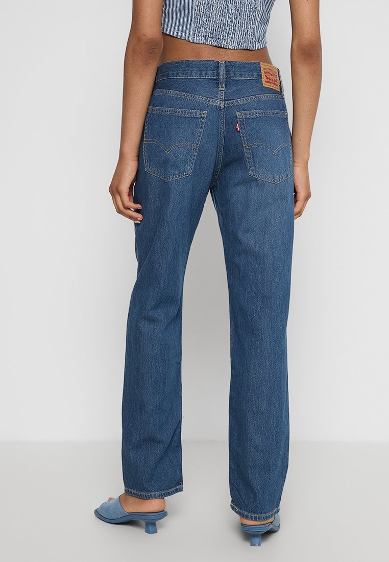 Femme portant un jean droit taille haute bleu, un top sans manches bleu et des mules à talons bleus, se tenant devant un fond uni.