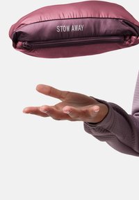 Hand in een paarse jas beschermende mouw die een opgevouwen, met een rits gesloten, roze tas met het label "STOW AWAY" tegen een lichte achtergrond houdt.