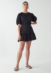 Czarna krótka sukienka z bufkowymi rękawami, dopasowaną talią i falbaniastym dołem. Gładki materiał z prostym wzorem. Noszona z cienkimi czarnymi sandałami.