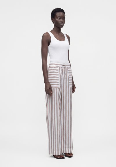 Γυναίκα στέκεται φορώντας λευκό αμάνικο κορμάκι με ριμπ ύφανση και φαρδιά μπεζ και λευκή κάθετα ριγέ παντελόνια με μεγάλες μπροστινές τσέπες.