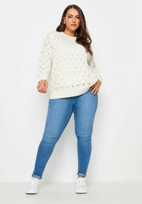 Elfenbeinfarbener, gestrickter Pullover mit Rautenmuster, Rundhalsausschnitt und langen Ärmeln, kombiniert mit hellblauen Skinny-Jeans und weißen Sneakers.
